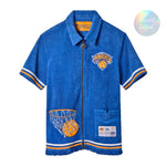 A bright blue terry cloth short-sleeve zip-up shirt with New York Knicks basketball details, including the Knicks logo, a basketball hoop patch on the lower side, and white-and-orange striped trim.