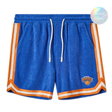 Bright blue terry cloth shorts with white drawstrings and orange-and-white stripes down the sides and hem, featuring the New York Knicks logo embroidered on one leg.