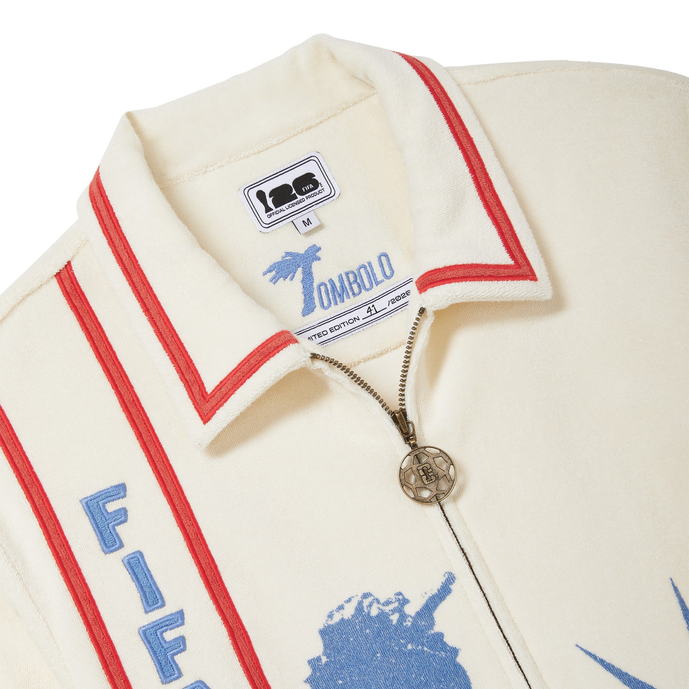 Close-up of beige cabana shirt collar with red piping, antique brass soccer zipper pull, and vertical FIFA World Cup 26 embroidered panel.