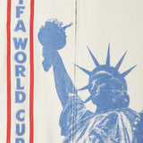 Close-up of beige cabana shirt showing vertical FIFA WORLD CUP embroidery beside large blue Statue of Liberty graphic.