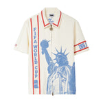 Beige short-sleeve zip-front cabana shirt with red contrast piping, vertical FIFA World Cup 26 lettering at wearer’s left, blue Statue of Liberty graphic across front, and 1951 armband detail on wearer’s right sleeve.