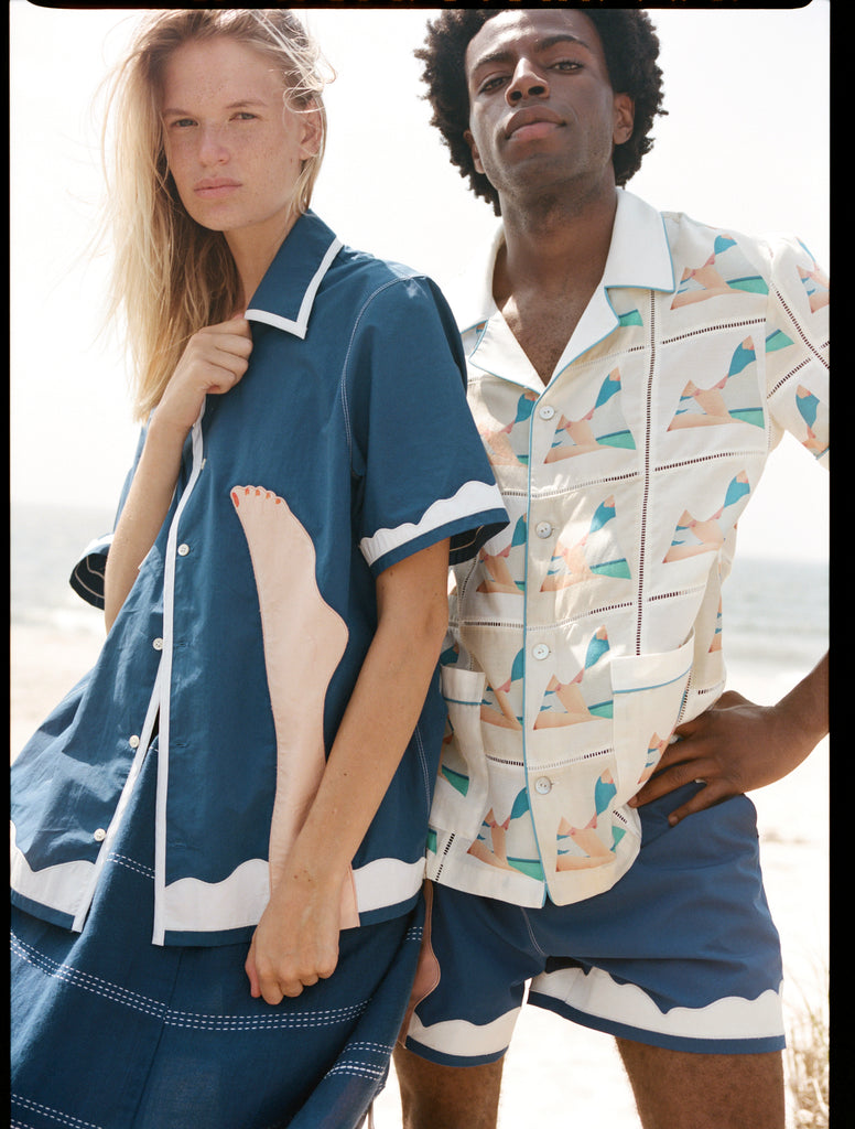 Woman holds closed unbuttoned blue seascape #2 shirt with man wearing matching trunks behind her