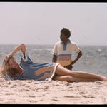Woman lies across beach with hand on her head while man gazes into the distance in background