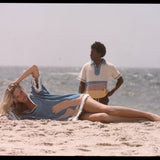 Woman lies across beach with hand on her head while man gazes into the distance in background