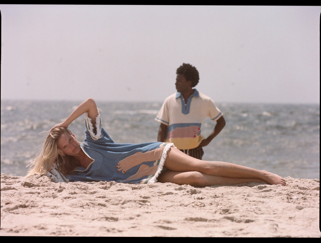 Woman lies across beach with hand on her head while man gazes into the distance in background