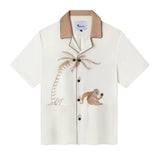 White hawaiian style button up featuring an embroidered monkey catching a falling coconut button with an embroidered palm tree arching into the placket which has buttons standing in for coconuts, also has lower patch pockets and is setailed with dark brown piping and a tan contrast collar
