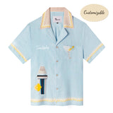 A light blue linen cabana shirt with cocktail-themed embroidery, including a cocktail shaker and a martini glass, and yellow Rick rack trim. The shirt is customized with 'Tombolo' on the chest. 