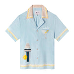 A light blue linen cabana shirt with cocktail-themed embroidery, including a cocktail shaker and a martini glass, and yellow Rick rack trim. The shirt is customized with 'Tombolo' over the pocket. 