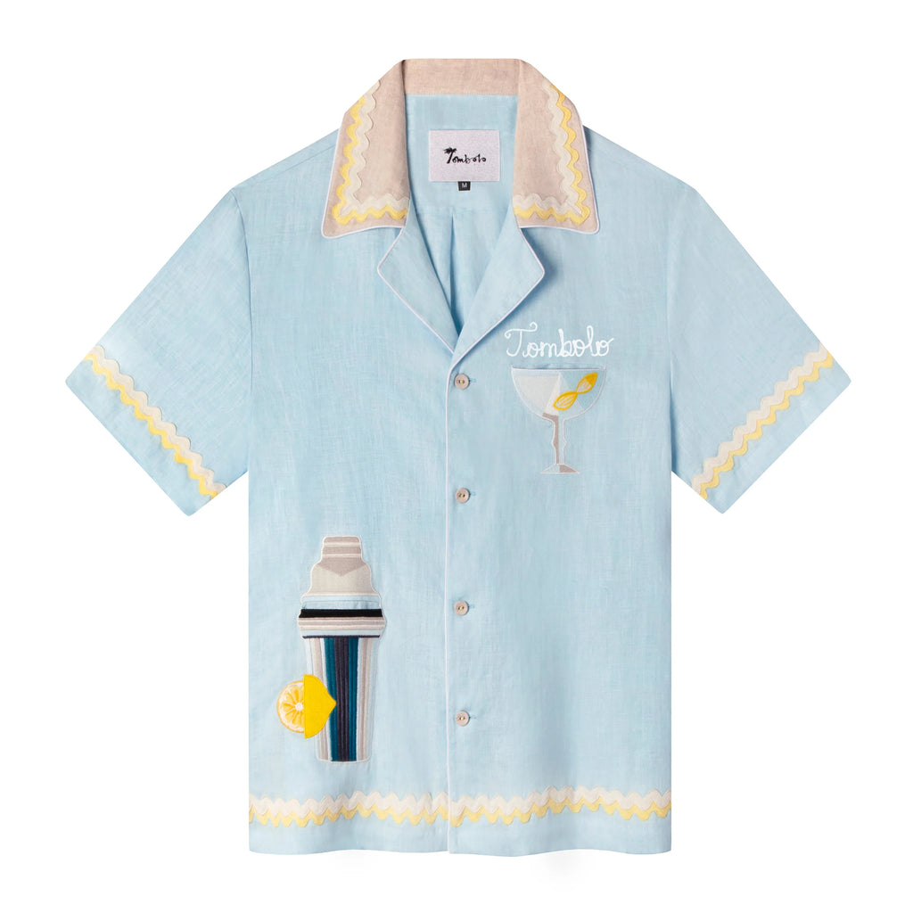 A light blue linen cabana shirt with cocktail-themed embroidery, including a cocktail shaker and a martini glass, and yellow Rick rack trim. The shirt is customized with 'Tombolo' over the pocket. 