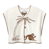 A cream-colored, sleeveless terrycloth top with a relaxed fit and brown zigzag trim along the edges. The front features an embroidered brown palm tree stretching across the chest, and a playful brown monkey reclining near the hem, holding a coconut. The shirt fastens with two visible brown buttons at the center. 