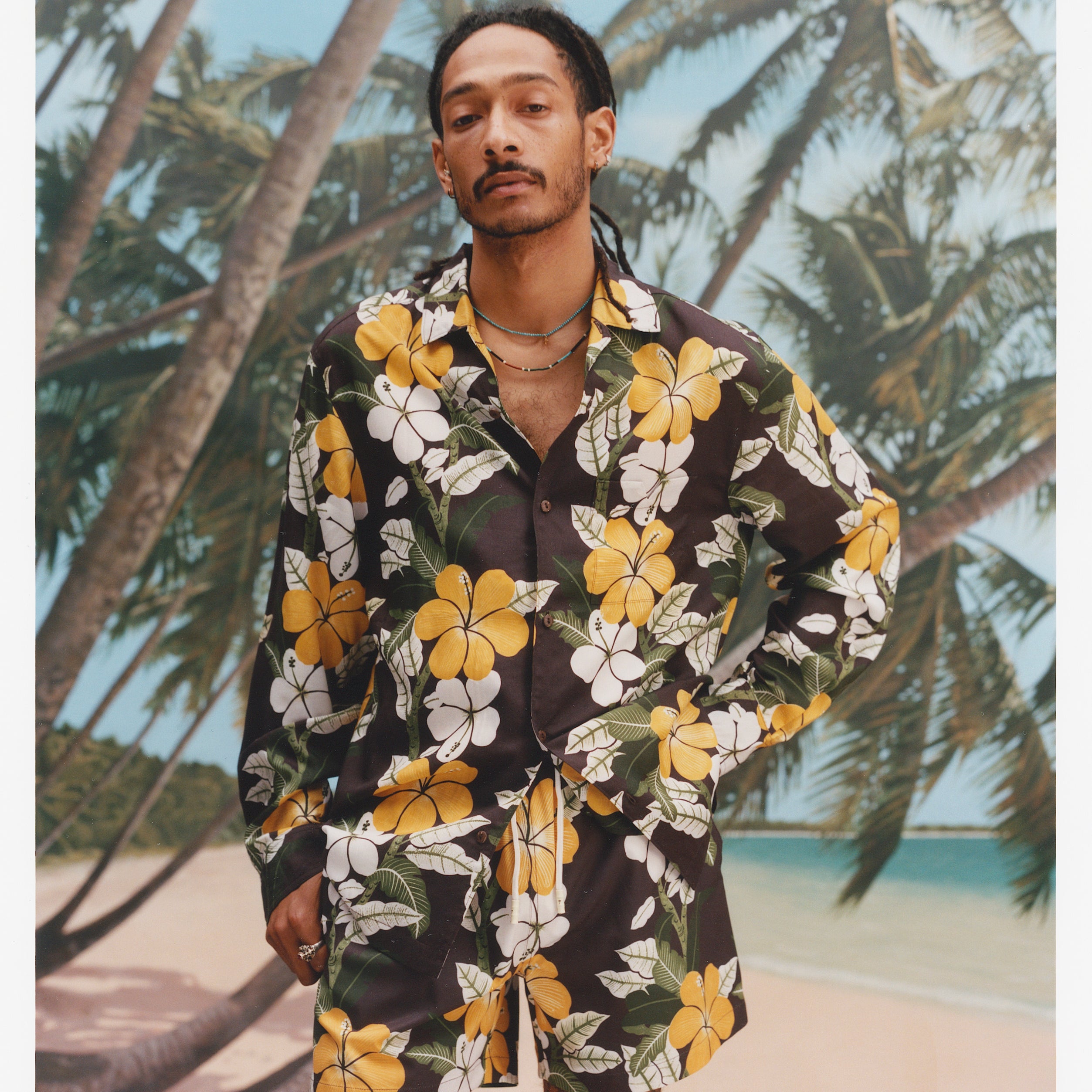 young male model wearing "Night Bloom" long-sleeved buttoned shirt with one button undone that features brown base and hibiscus pattern alongside matching swim trunks with a brown base and hibiscus pattern with a background of leaning palm trees on quiet beach.