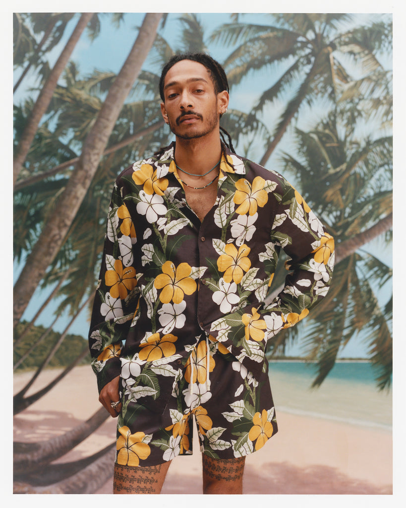 young male model wearing "Night Bloom" long-sleeved buttoned shirt with one button undone that features brown base and hibiscus pattern alongside matching swim trunks with a brown base and hibiscus pattern with a background of leaning palm trees on quiet beach.