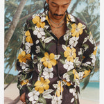 Closer up image of a young male model wearing the "Night Bloom" long-sleeve shirt with one button undone and one hand in his pocket with a background of palm trees and a beach. 