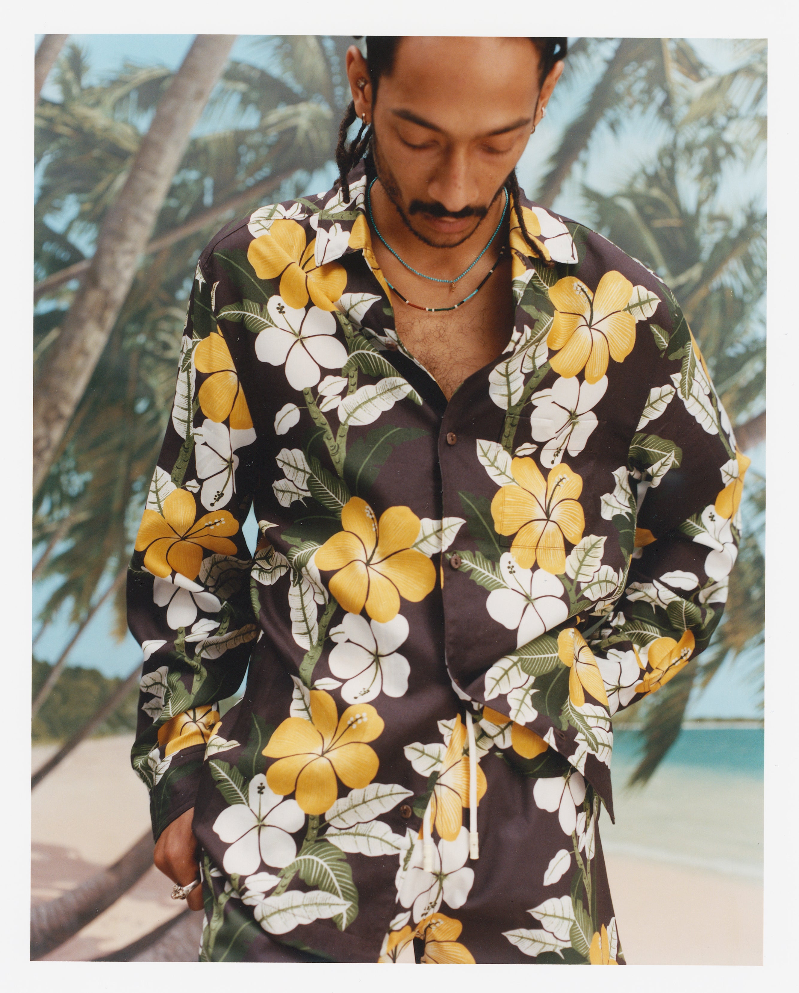 Closer up image of a young male model wearing the "Night Bloom" long-sleeve shirt with one button undone and one hand in his pocket with a background of palm trees and a beach. 