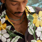Young male model wearing "Night Bloom" long-sleeve shirt with one button undone that showcases the curved cuff and placket on the shirt and hibiscus pattern while also wearing a teal colored beaded necklace.