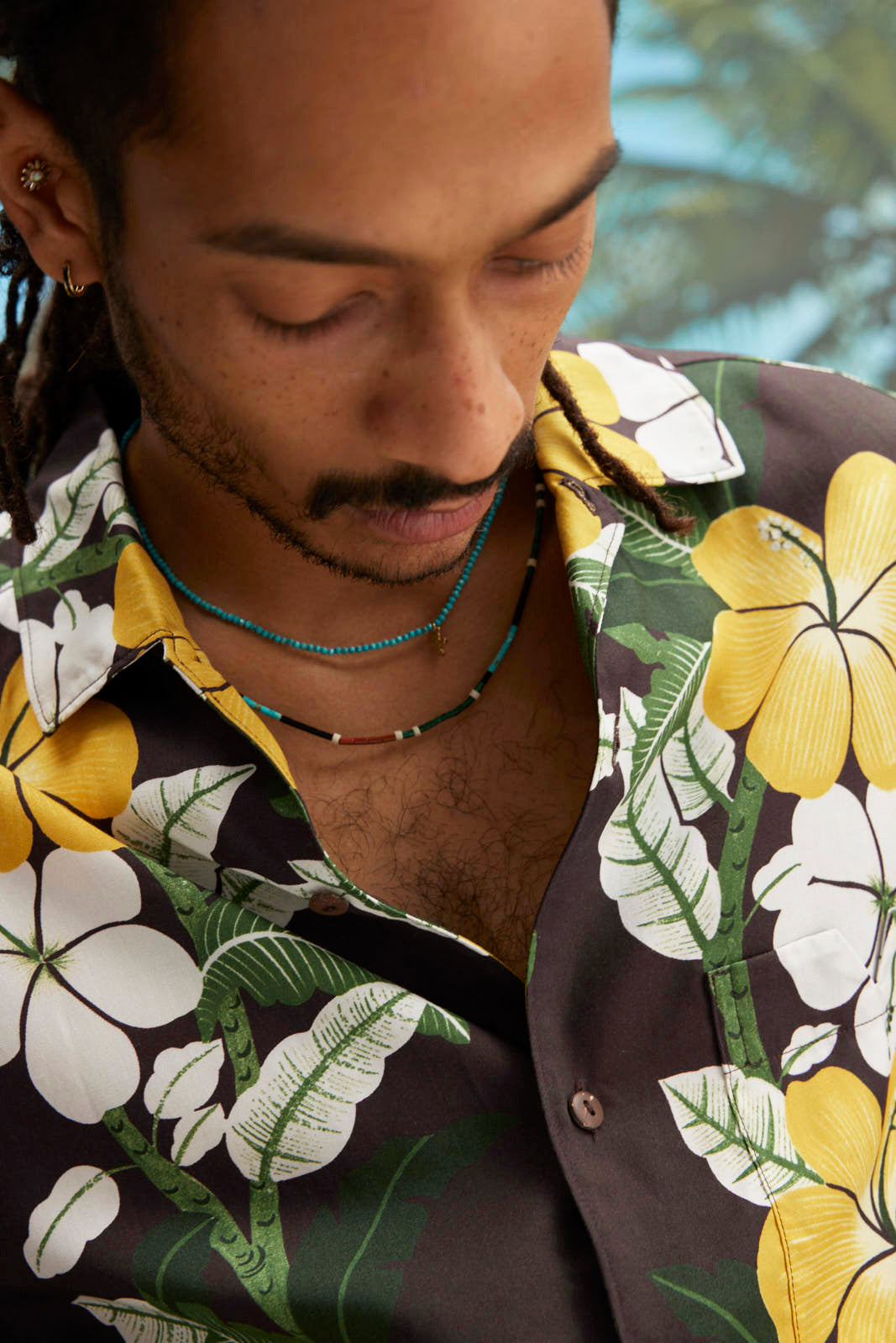 Young male model wearing "Night Bloom" long-sleeve shirt with one button undone that showcases the curved cuff and placket on the shirt and hibiscus pattern while also wearing a teal colored beaded necklace.