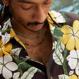 Young male model wearing "Night Bloom" long-sleeve shirt with one button undone that showcases the curved cuff and placket on the shirt and hibiscus pattern while also wearing a teal colored beaded necklace.