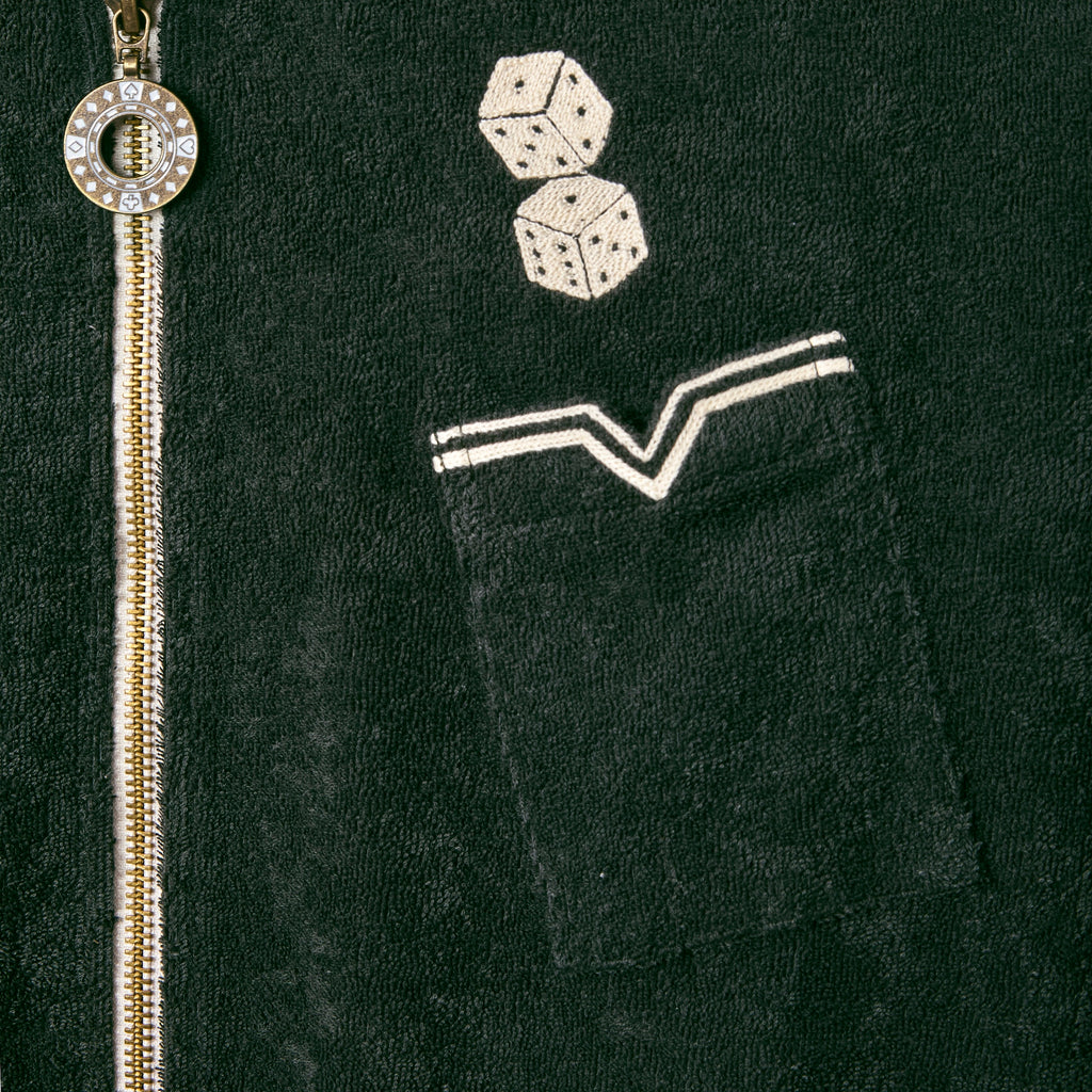 Close-up of a dark green terry cloth shirt showing a small chest pocket with white piped trim and two embroidered dice above it. A silver zipper with a circular pull tab resembling a poker chip runs vertically beside the pocket.