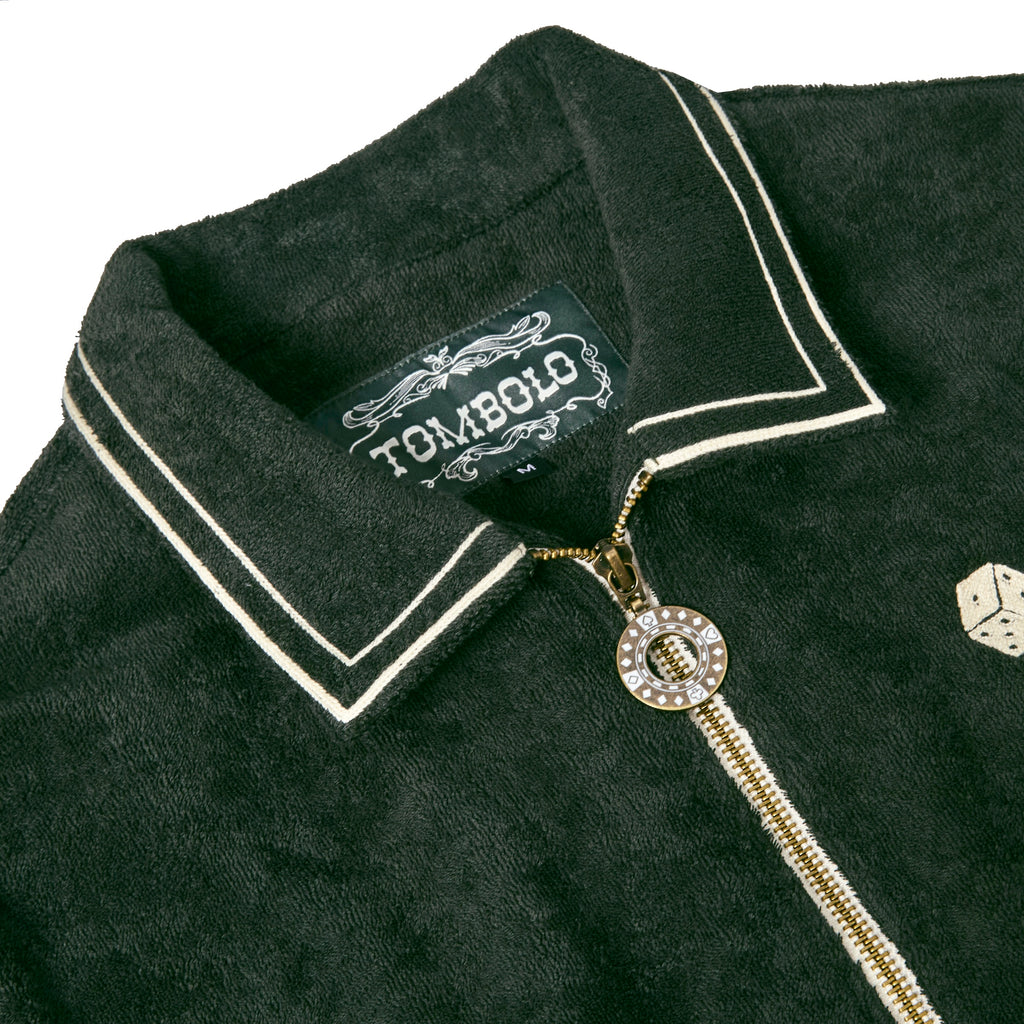 Close-up of the collar and zipper of a dark green terry cloth shirt with white piping. A decorative gold zipper pull shaped like a poker chip is partially unzipped beneath a black woven label that reads “TOMBOLO” in white vintage-style lettering. The edge of an embroidered dice graphic is visible to the right.