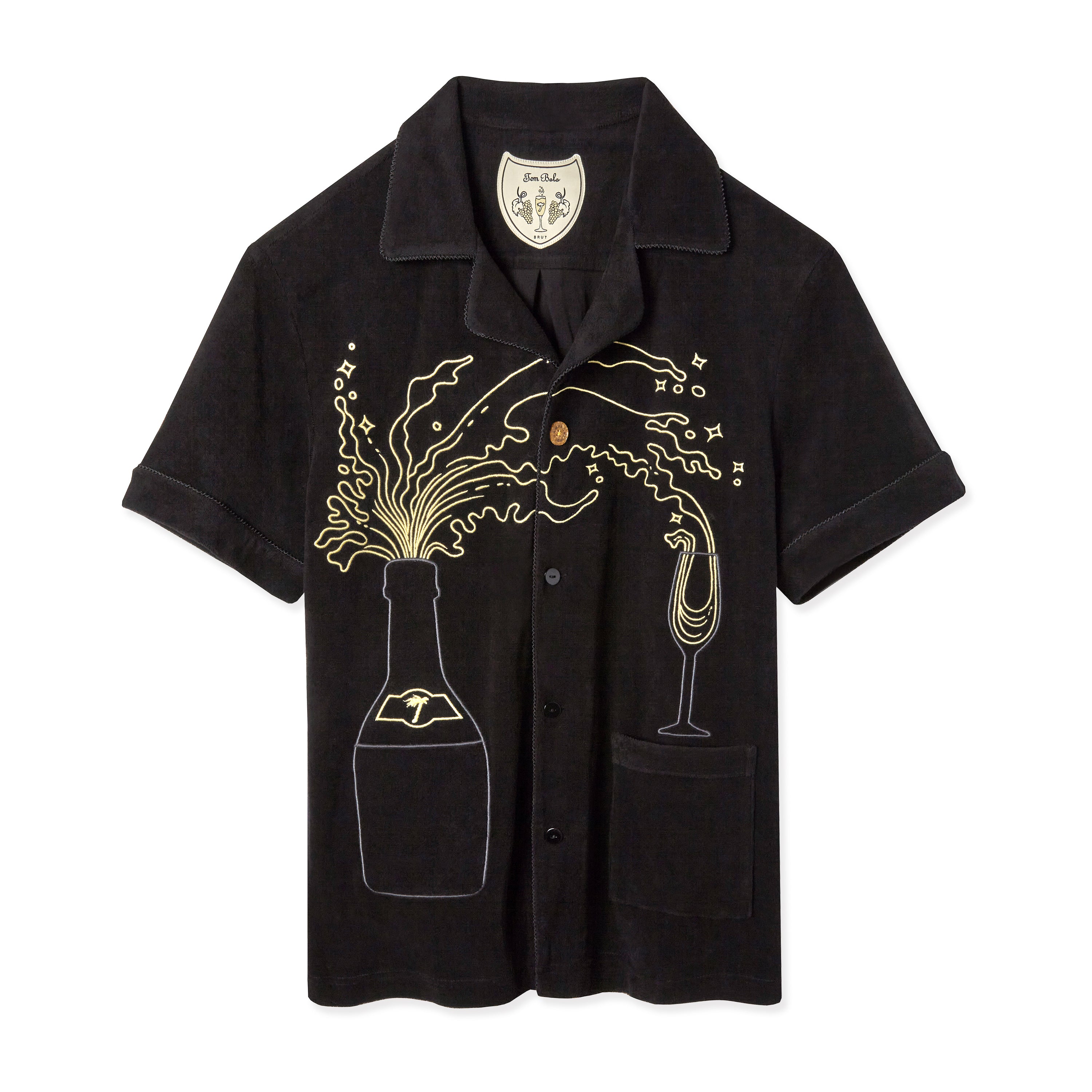 Black short-sleeve button-up shirt with playful embroidery depicting a champagne bottle popping open, with golden embroidered fizz and bubbles bursting across the chest. A champagne flute on the right side catches the stream. The shirt features a chest pocket and a soft, textured fabric with a relaxed fit.