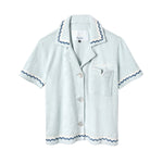 Light blue terrycloth crop-cut shirt with short sleeves and scalloped trim in navy and white. Features a front chest pocket with flap, white embroidered 'Tombolo' text on the chest, and mother-of-pearl buttons.