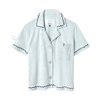 Light blue terrycloth crop-cut shirt with short sleeves and scalloped trim in navy and white. Features a front chest pocket with flap, white embroidered 'Tombolo' text on the sleeve, and mother-of-pearl buttons.