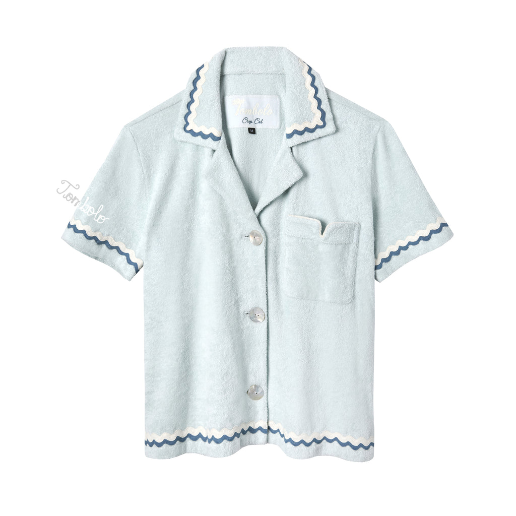 Light blue terrycloth crop-cut shirt with short sleeves and scalloped trim in navy and white. Features a front chest pocket with flap, white embroidered 'Tombolo' text on the sleeve, and mother-of-pearl buttons.