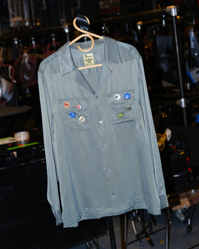 A green button-up shirt with multiple colored buttons on the chest, adorned with 'SNL' themed pins.
