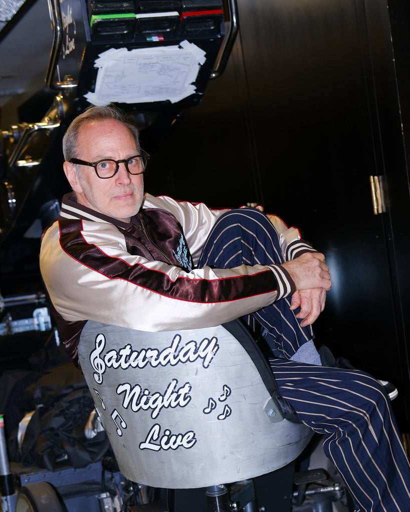SNL costume designer, Tom Broecker, is seen sitting in the SNL studio, while wearing the 'SNL" Souvenir Jacket and Navy Easy Going Striped pants. 