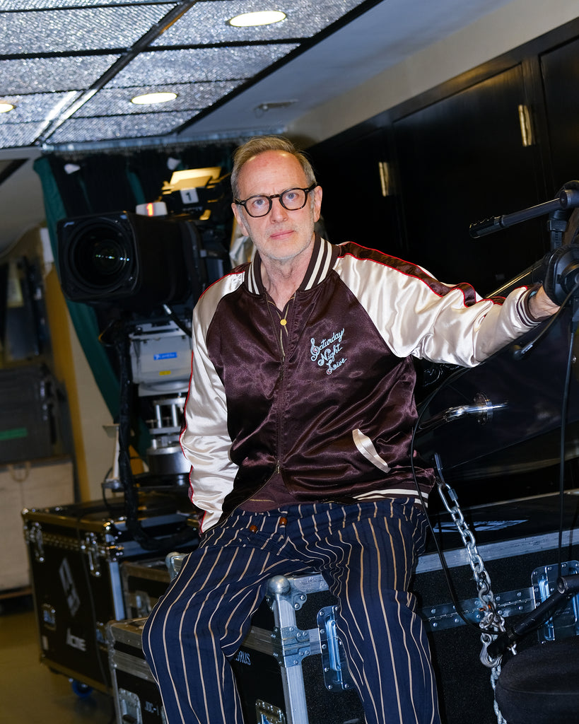 SNL costume designer, Tom Broecker, is seen sitting in the SNL studio, while wearing the 'SNL" Souvenir Jacket and Navy Easy Going Striped pants. 