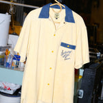 A pale yellow terry cloth short-sleeved shirt with mid-wash denim details, featuring a 'Saturday Night Live' logo on the chest pocket and custom snap buttons, hanging at the SNL studio.