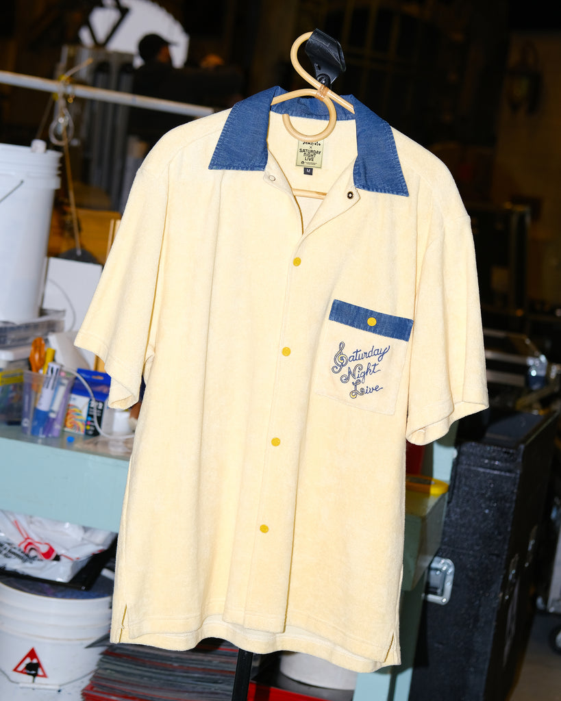 A pale yellow terry cloth short-sleeved shirt with mid-wash denim details, featuring a 'Saturday Night Live' logo on the chest pocket and custom snap buttons, hanging at the SNL studio.