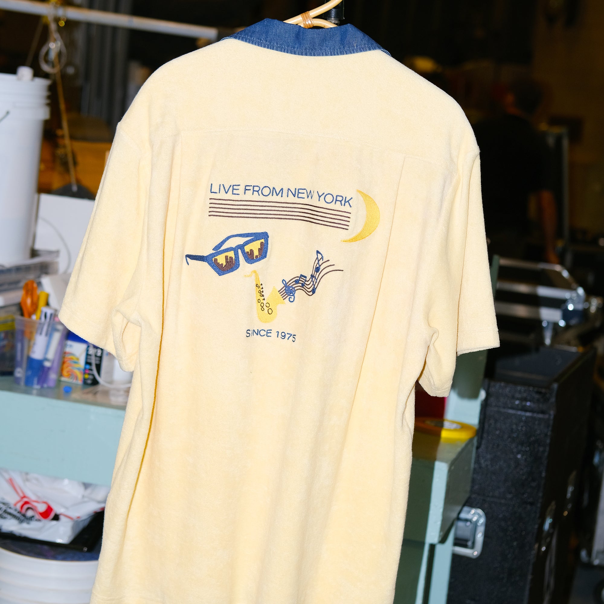 Backside of pale yellow terry cloth cabana shirt, featuring original artwork chain-stitched on the back, paying homage to SNL’s rich history of musical guests and the promise of a crescent moon rising over the greatest city in the world on a Saturday night.