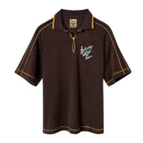 A brown Saturday Night Live x Tombolo polo shirt with yellow overlock stitching and a chain-stitched 'Saturday Night Live' logo on the left chest.