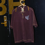 A brown Saturday Night Live x Tombolo polo shirt with yellow overlock stitching and a chain-stitched 'Saturday Night Live' logo on the left chest, hanging on a hanger at the SNL studio.