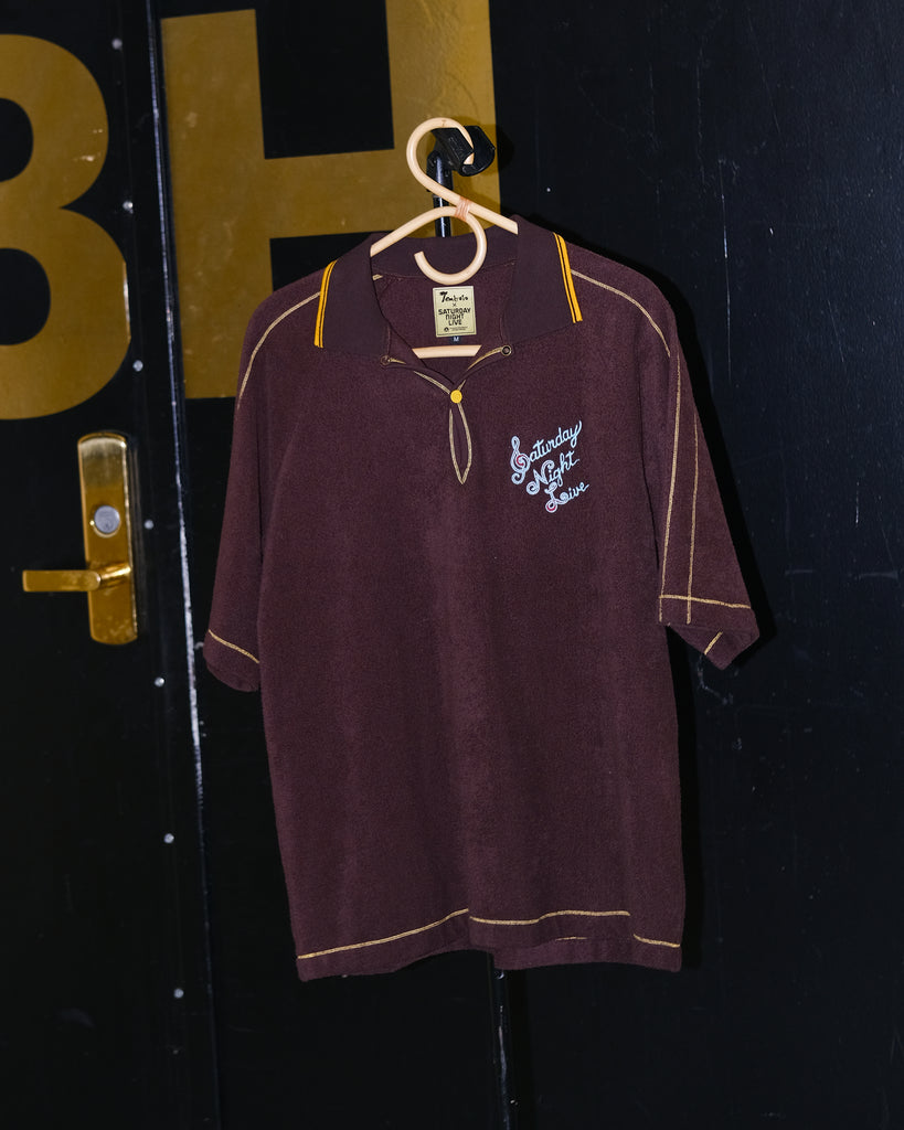 A brown Saturday Night Live x Tombolo polo shirt with yellow overlock stitching and a chain-stitched 'Saturday Night Live' logo on the left chest, hanging on a hanger at the SNL studio.