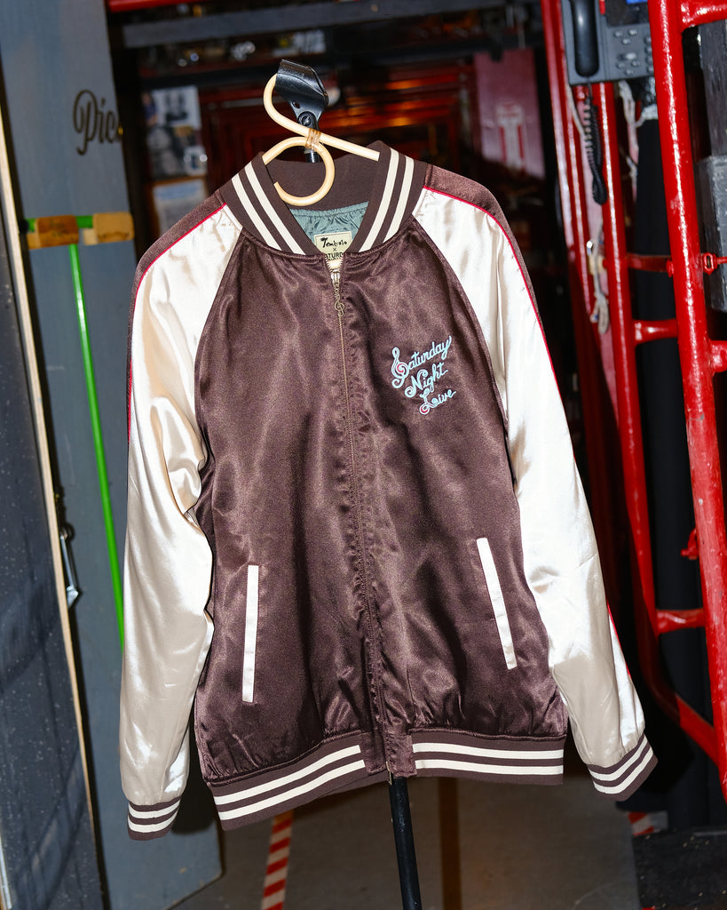 A satin souvenir jacket with 'Saturday Night Live' text and design elements, featuring a mix of maroon, beige, and white colors with red piping.