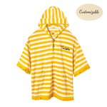 Yellow and white striped terrycloth poncho with short sleeves and yellow fringe trim. Features a hood with a tie closure, front pouch pocket, and black embroidered 'Tombolo' text on the chest.