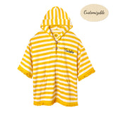 Yellow and white striped terrycloth poncho with short sleeves and yellow fringe trim. Features a hood with a tie closure, front pouch pocket, and black embroidered 'Tombolo' text on the chest.