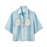 Light blue, short-sleeve button-up shirt with a relaxed fit. The shirt features an embroidered design resembling a sand dollar bikini top, with two large cream-colored sand dollars at the chest and shell bead strands extending from the shoulders, across the chest, and down the sides.