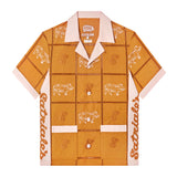 Front-facing product image of 'The Sopranos' Satriale's (Pruh-Zhoot) that is a mustardy-orange color and features square panels joined by ‘ladder’ lace embroidery, piping on cuffs and pocket strips, custom woven 'The Sopranos' x Tombolo interior label, including special pig logo from Satriale's storefront, matching poplin-wrapped buttons, chain-stitched Satriale's logo on side panels, and pig embroidery on alternating panels, labeled with various cuts in Sopranos parlance: "gabagool", "pruh-zhoot", etc.