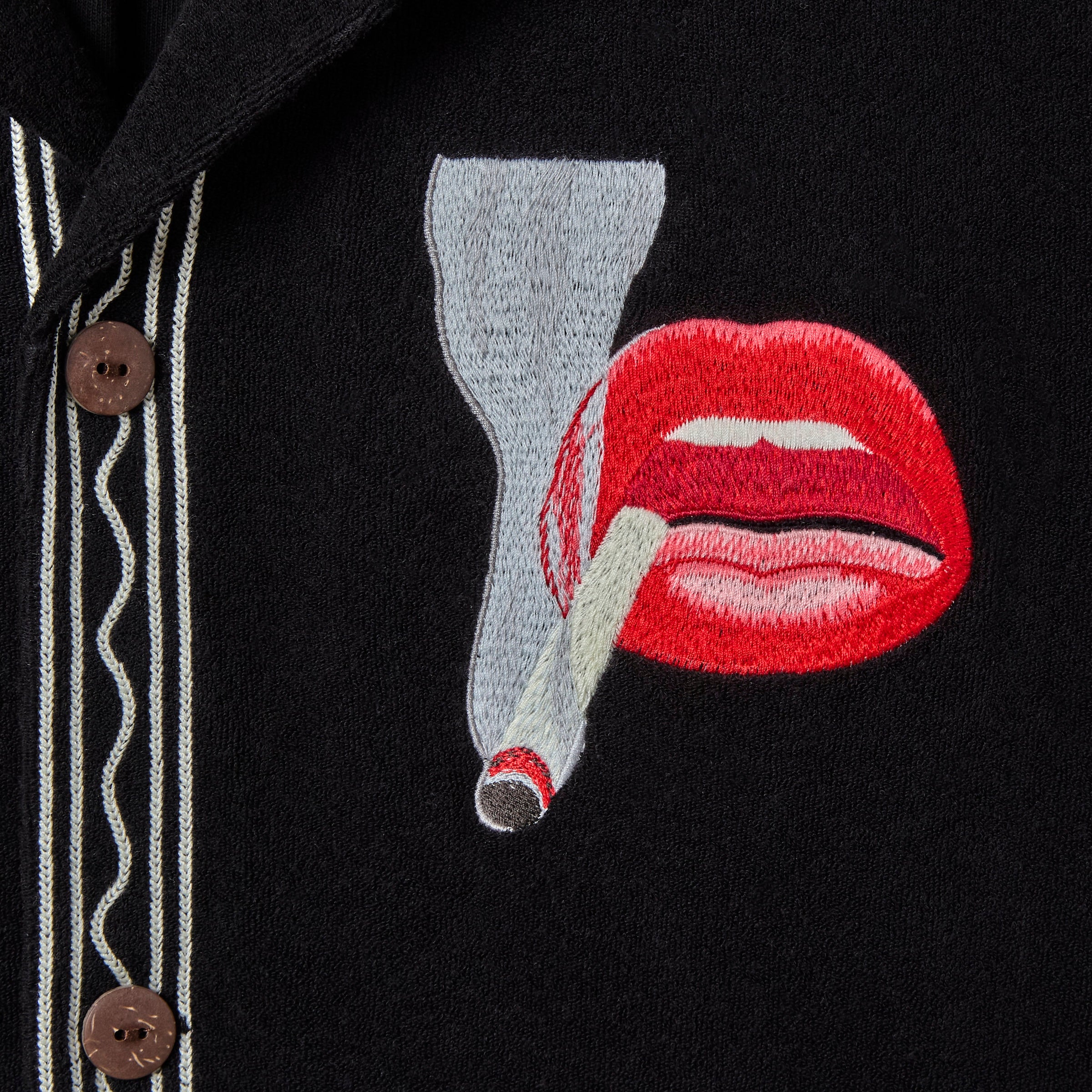 Close-up of the chest embroidery on a black terry cloth shirt featuring red lips holding a lit cigarette, with contrast piping and brown button detail nearby.