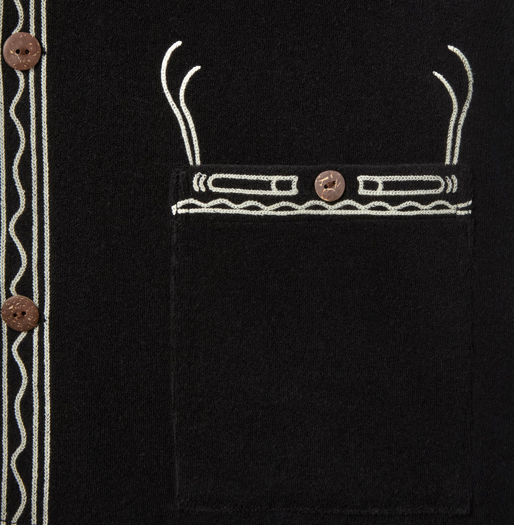 Close-up of a black terry cloth pocket with off-white embroidered trim and a brown button. The stitching resembles a stylized cigarette motif with curling smoke lines above.