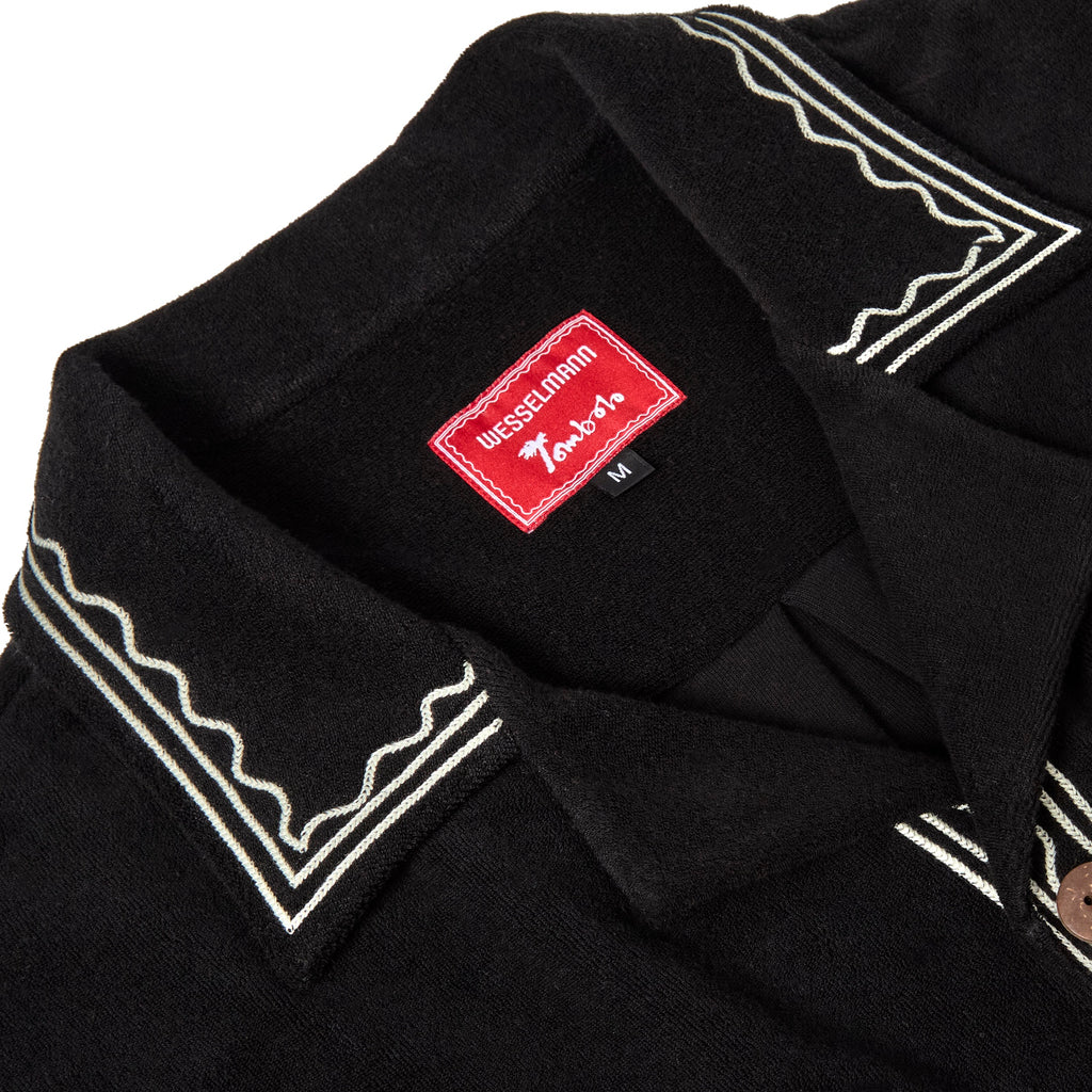 Close-up of the collar on a black terry cloth shirt with cream embroidered trim and a red woven label that reads 'Wesselmann Tombolo'.