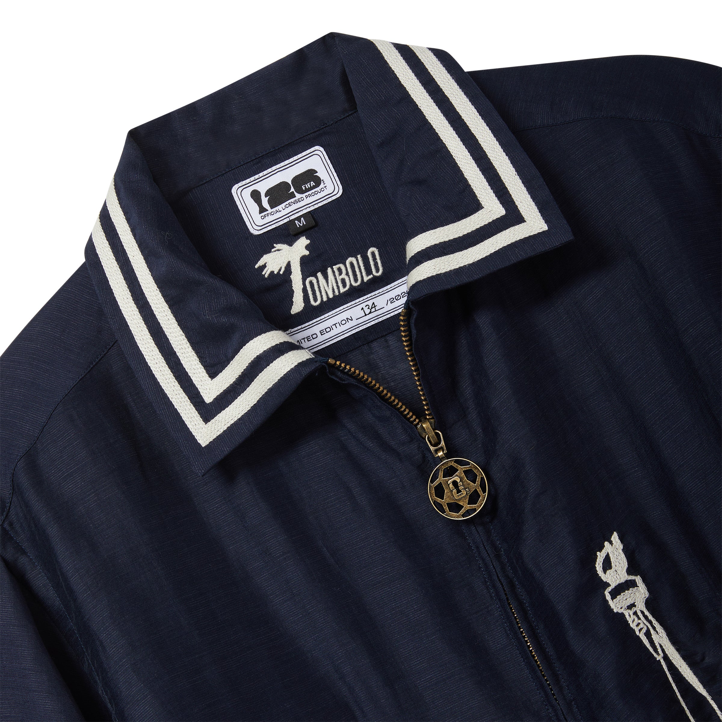 Close-up of navy cabana shirt collar with white striped trim, full-zip closure, and antique brass soccer-ball zipper pull.