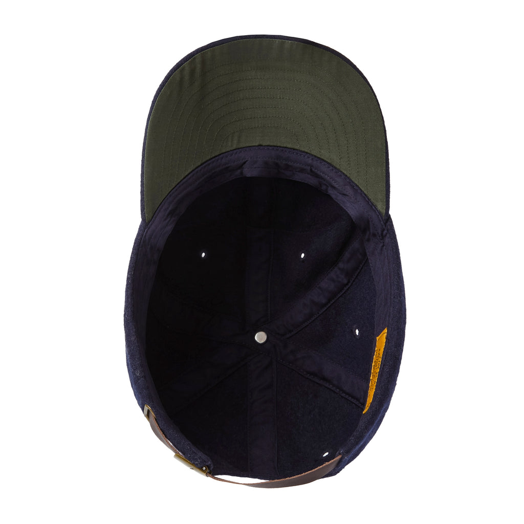 Interior view of a navy wool baseball cap with a green underside on the brim. The crown features six panels with stitched eyelets for ventilation and a central button at the top. A brown leather strap with brass hardware is partially visible at the back for adjustable sizing.