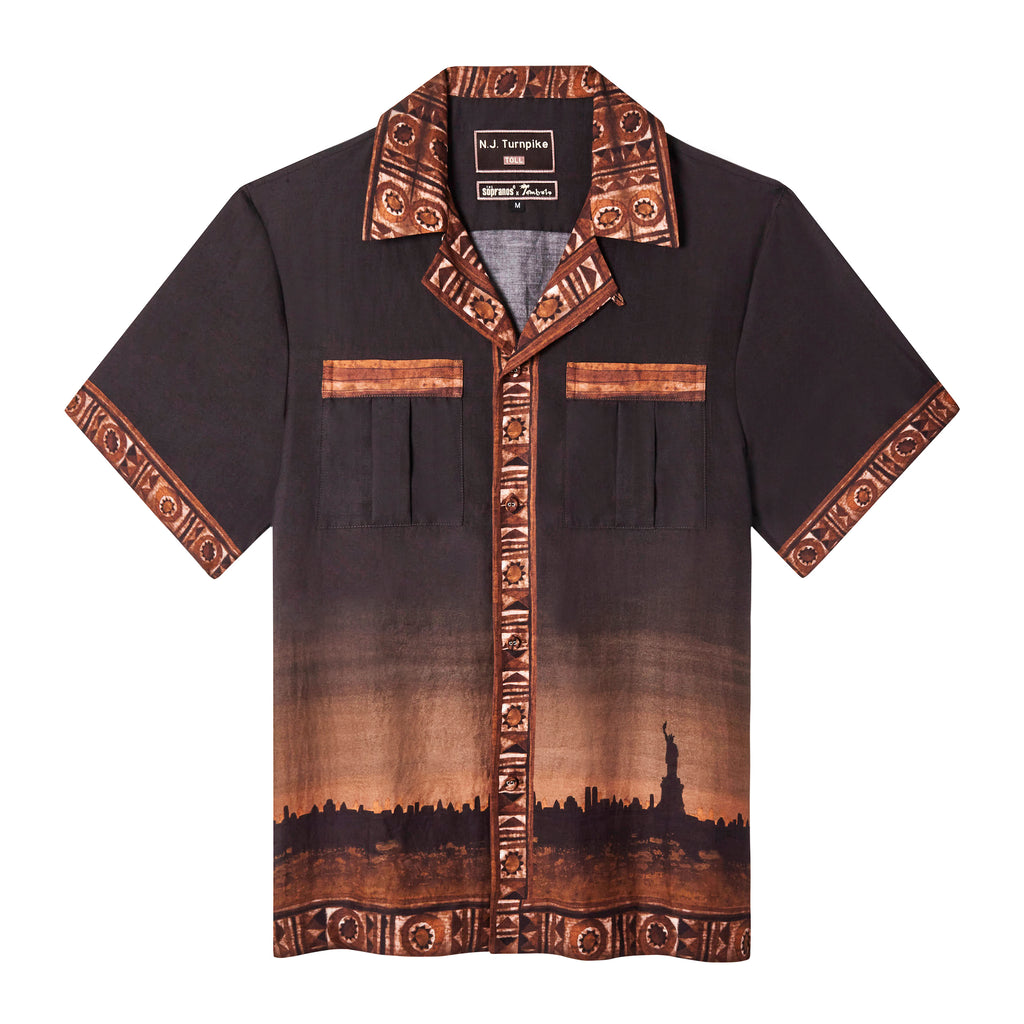 Front-facing product image of the 'The Sopranos' Turnpike Batik that features a digitally printed skyline of New York City including the Statue of Liberty, a custom woven 'The Sopranos' x Tombolo' collaboration label, and finally a contrasting batik collar, cuffs, placket, and bottom sweep, all of which exists on a brown gradient that resmebles a setting sun over the cityscape. Shot on a white background.