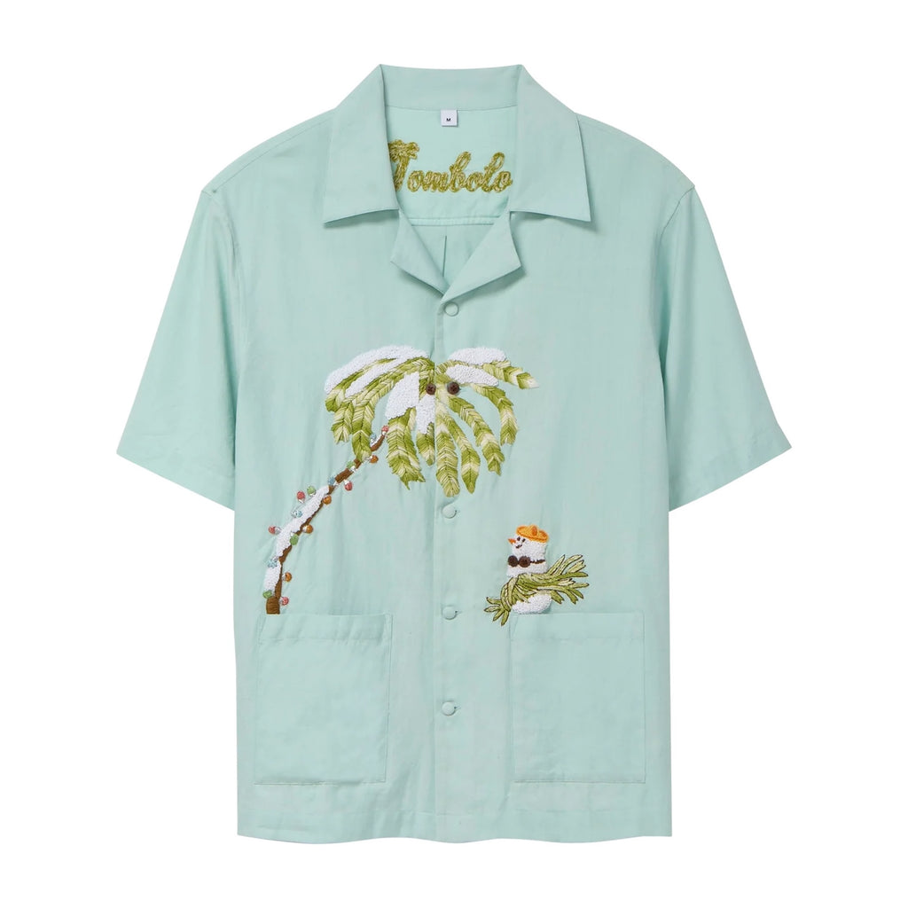 Light blue short-sleeve cabana shirt featuring embroidered artwork of a snowy palm tree and a small snowman with sunglasses and a drink. The shirt has two front patch pockets and a chain-stitched “Tombolo” logo at the collar.