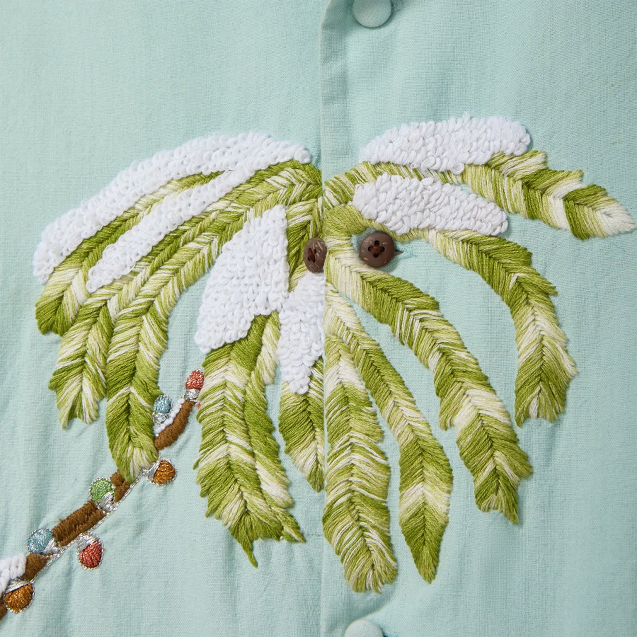 Close-up of a light blue cabana shirt showing embroidered palm tree fronds with white snowy accents and decorative multicolored lights along the trunk.