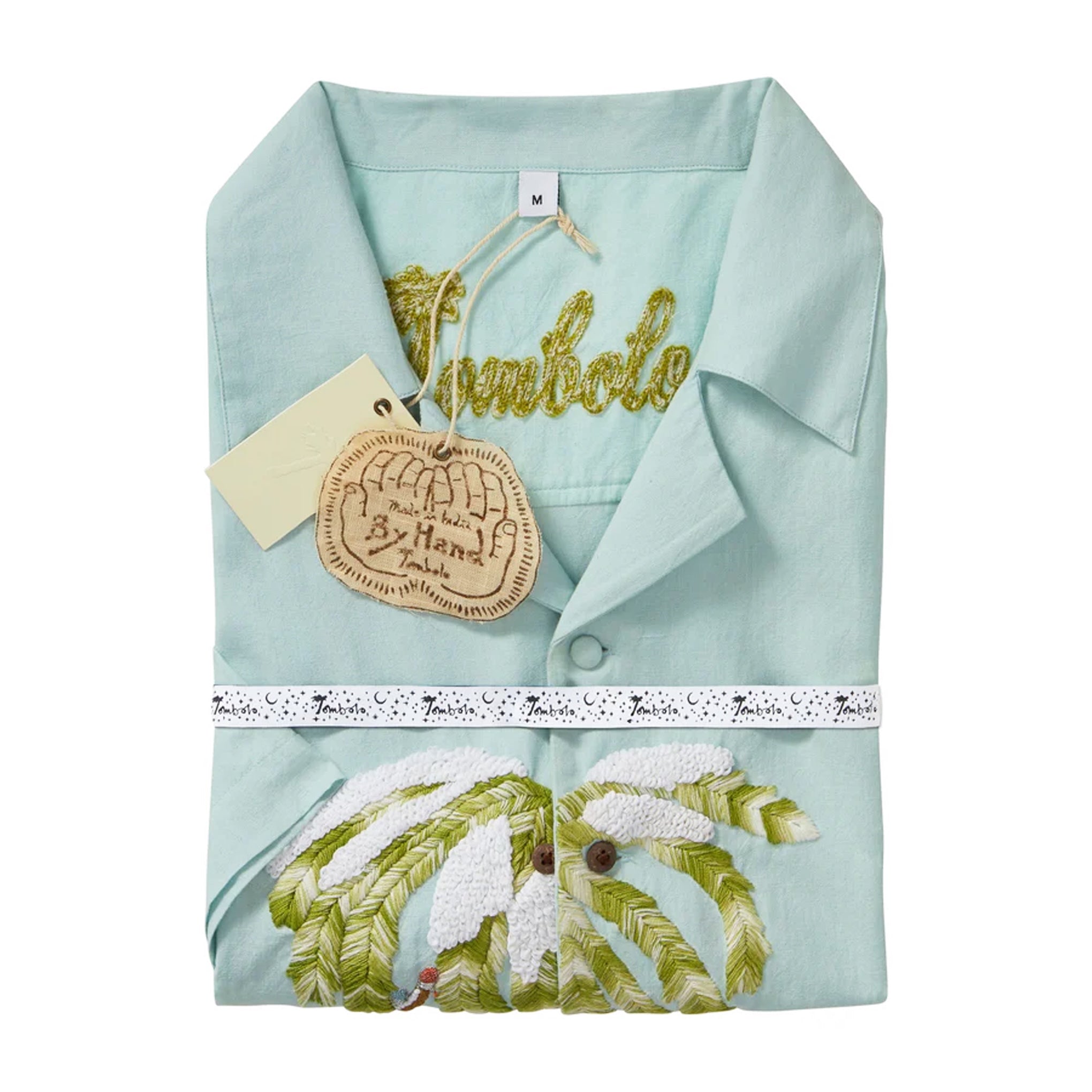Folded light blue cabana shirt with embroidered snowy palm tree detail, shown with a chain-stitched “Tombolo” logo at the collar. The shirt is wrapped with branded ribbon and includes attached hangtags.